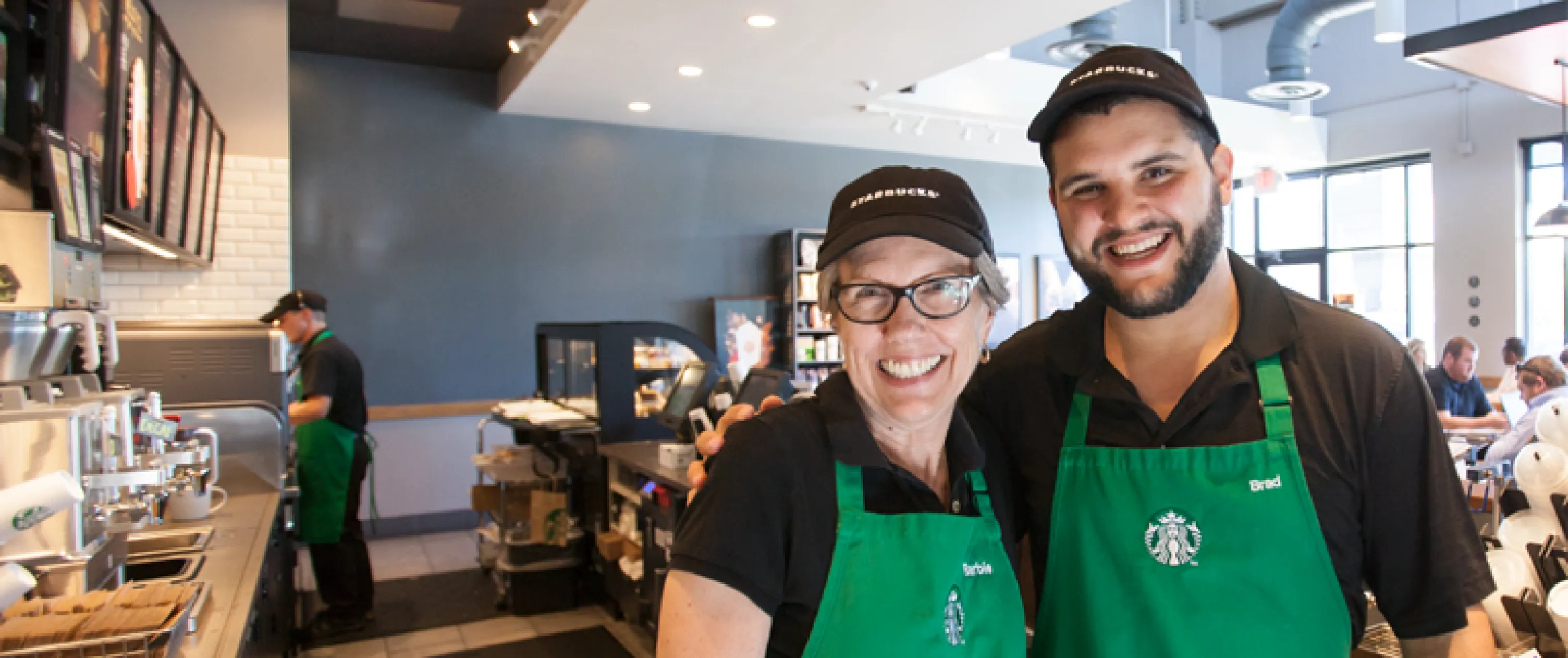 2 Starbucks partners (employees) proudly wearing their green aprons and starbucks caps.