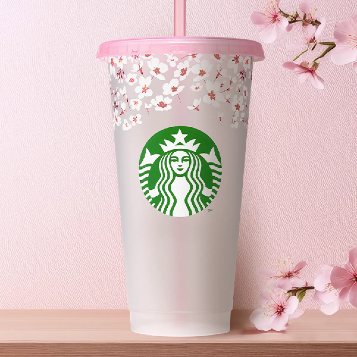 A clear plastic Reusable Cold Cup, decorated with pale pink Sakura and a translucent pink lid and straw, holds 24oz, on a tabletop with pink sakura background decoration