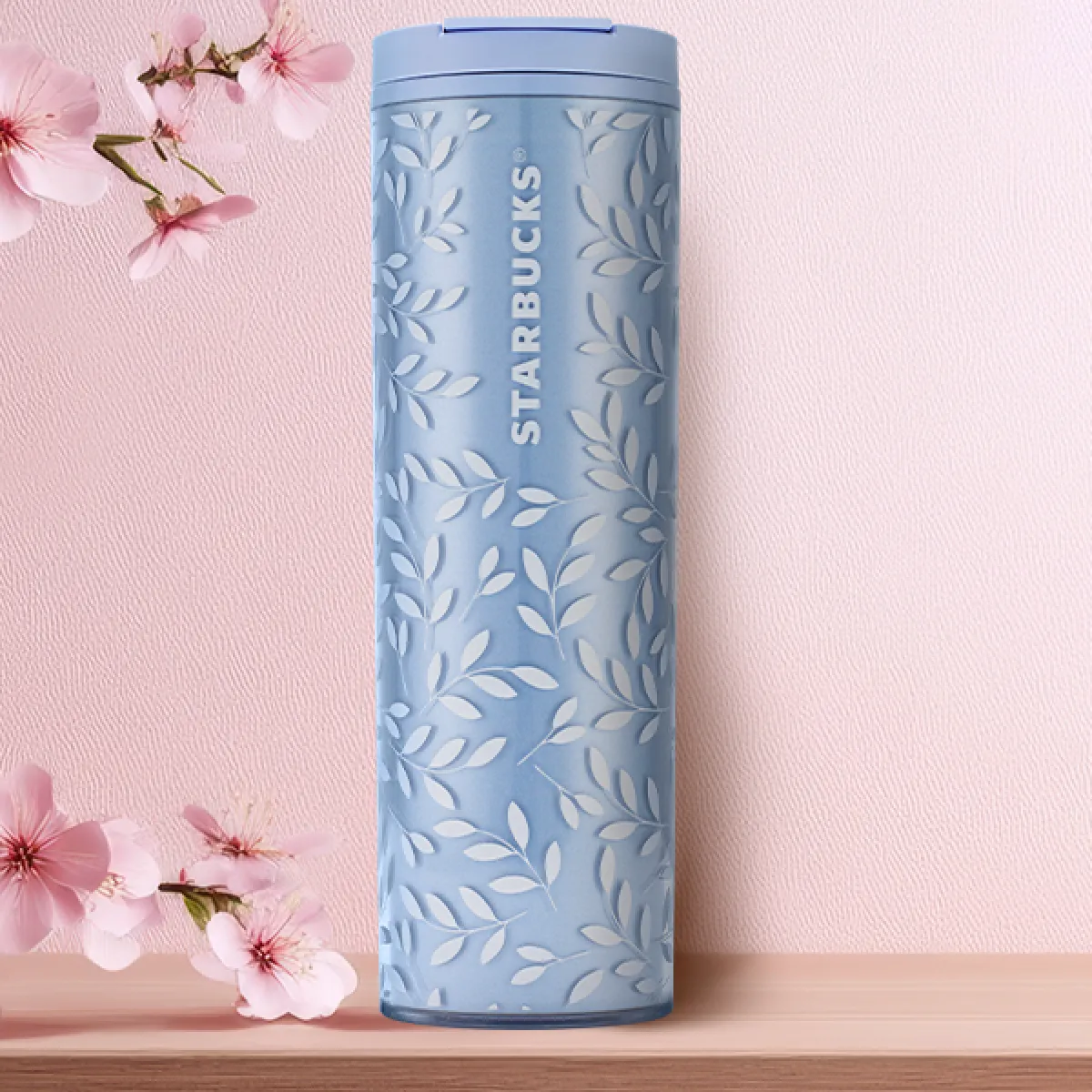 A light blue plastic tumbler, for hot drinks holding 16oz. Designed with a white petal 'Spring Breeze' design and a blue plastic flip-lid, on a tabletop with pale pink sakura decorated background.