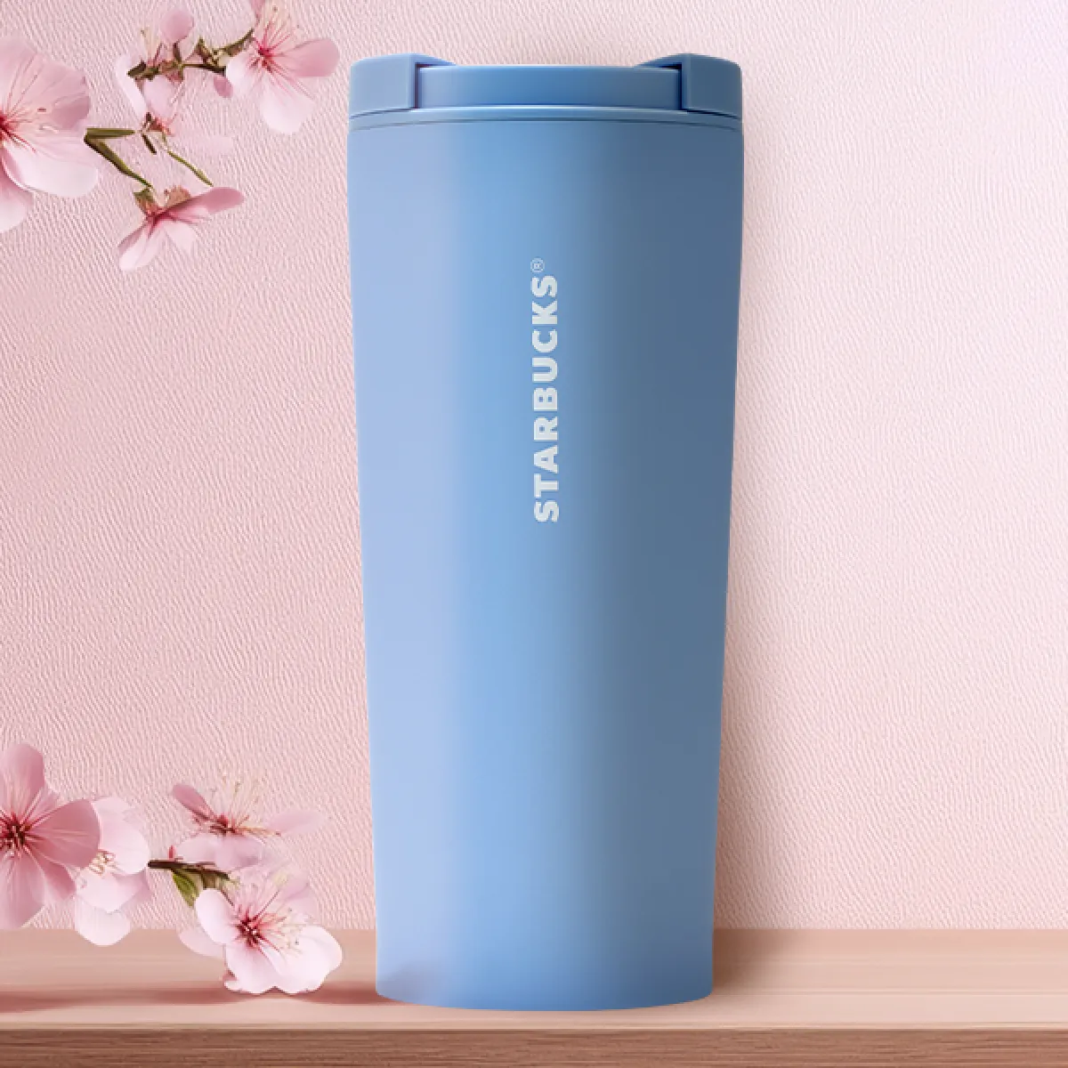 A cornflower blue 17oz tumbler, with a simple Starbucks logo design, and sleek twist-lid mechanic, on a tabletop with a pale pink sakura background design.