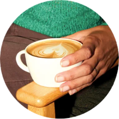 A photo of a hand holding a flat white in a mug, with heart-shaped latte art in the foam.