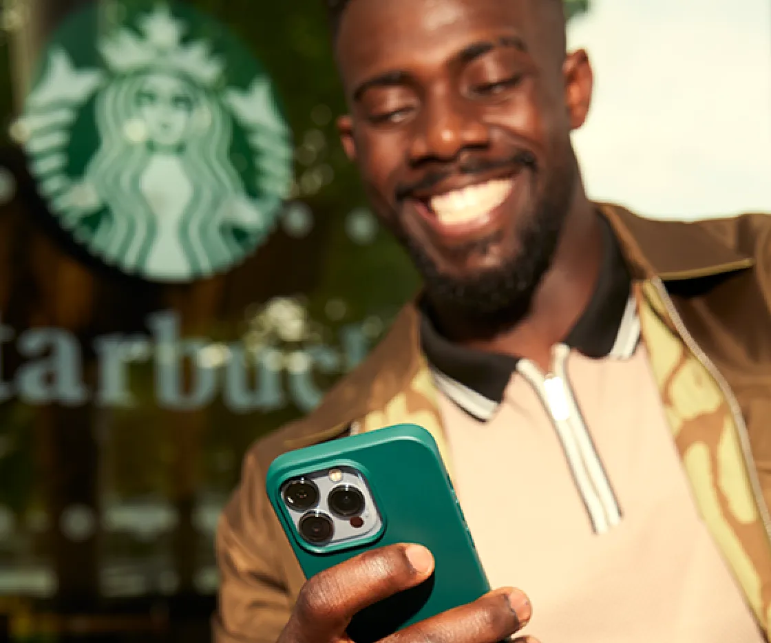 A person stands in front of a Starbucks® store with a phone in one hand, smiling.