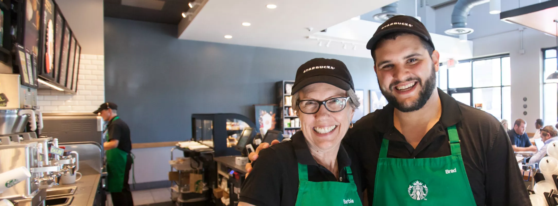 2 Starbucks partners (employees) proudly wearing their green aprons and starbucks caps.