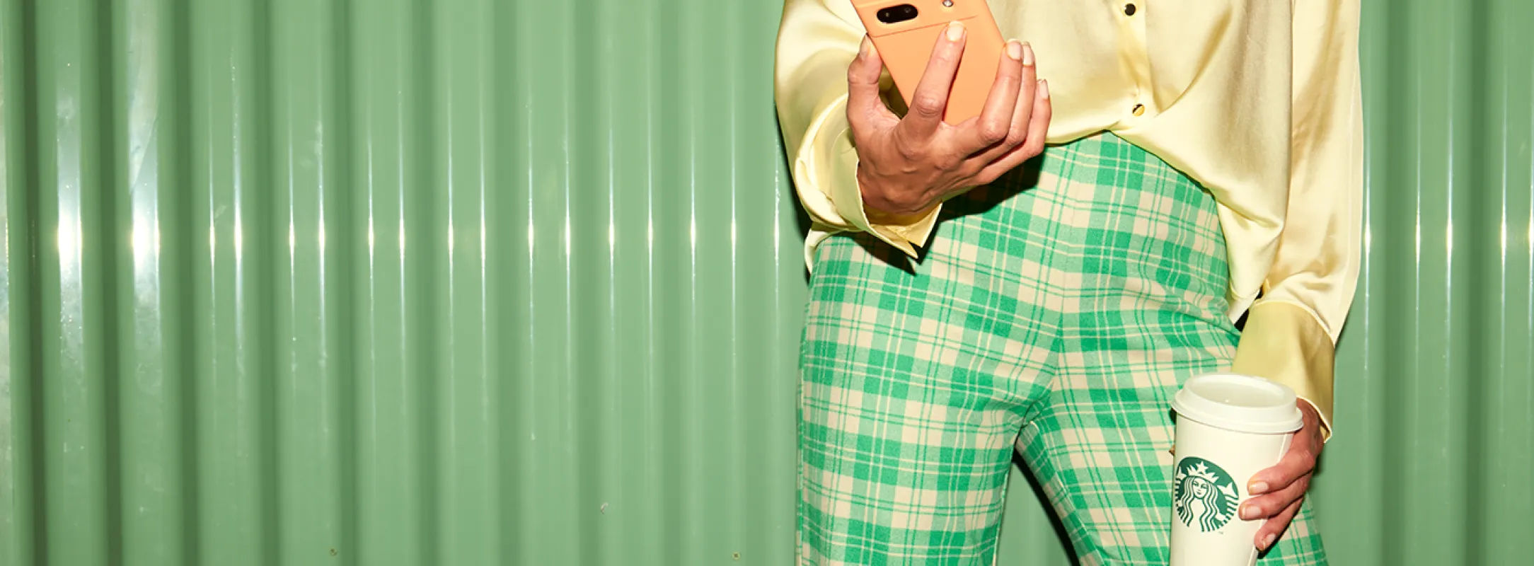 A person stands against a green wall with a phone in one hand, and a Starbucks in the other