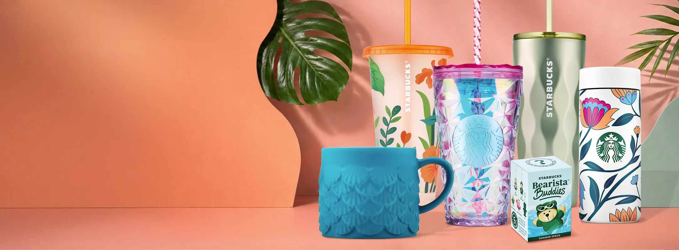 A handful of our merchandise collection against a peachy summer background table, with tropical leaves surrounding the collection. 