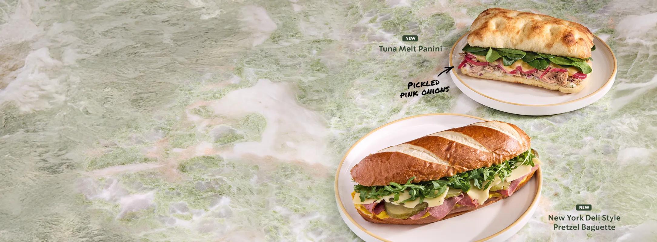 A photo of the new Tuna Melt Panini and New York Deli Style Pretzel Baguette on plates, showing their colourful ingredients with the product names next to them.