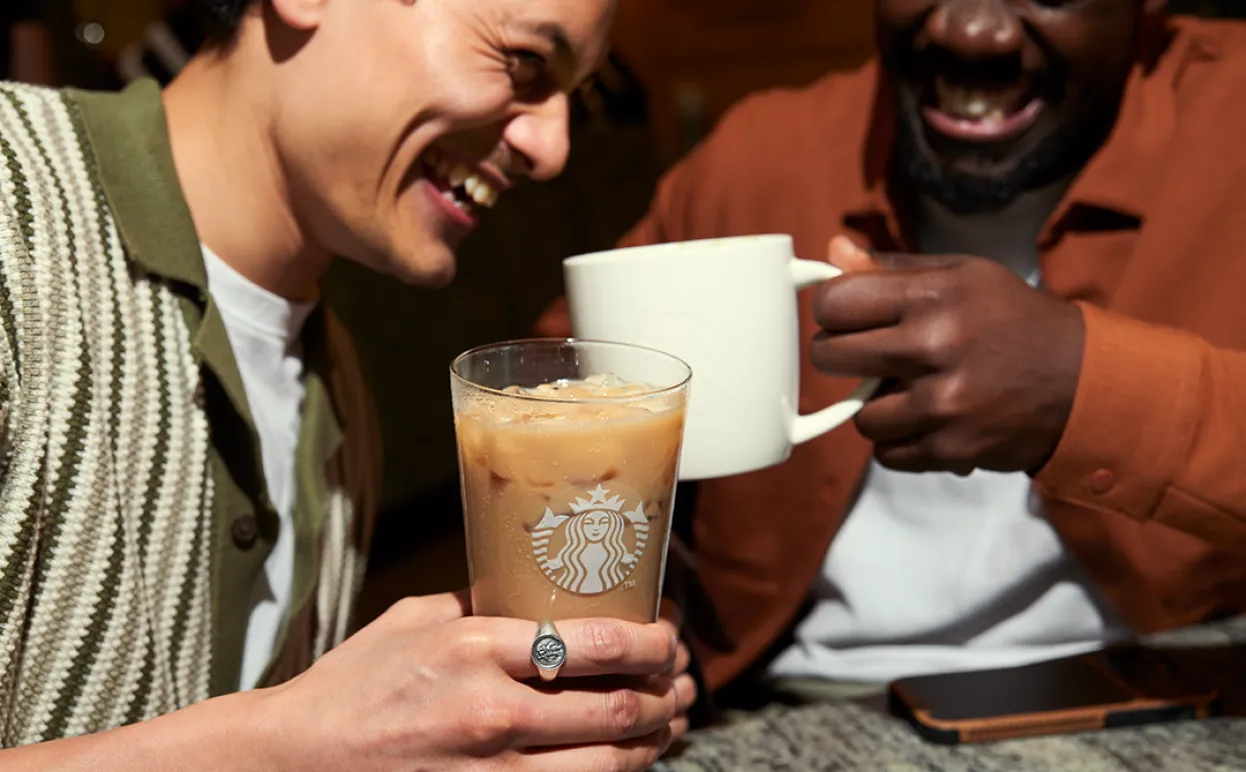 Two customers enjoy beverages whilst laughing together, one holds an iced drink in a glass whilst the other has a hot drink in classic Starbucks ceramic mug.