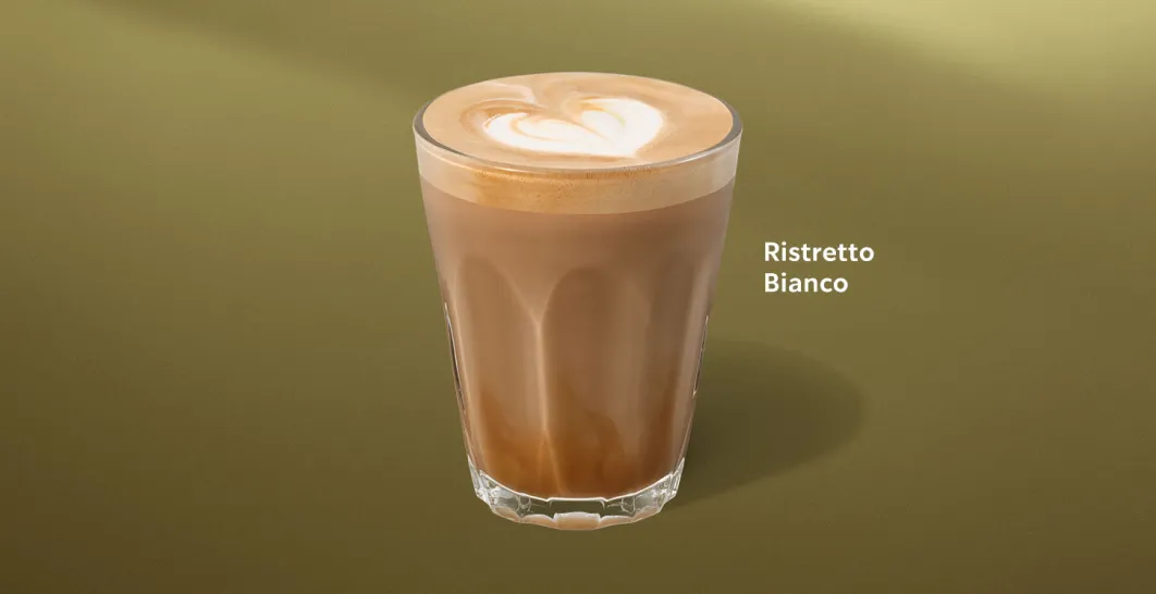 A photo of our new ristretto bianco - a short white coffee in a glass with heart shaped latte art, on a background of green.