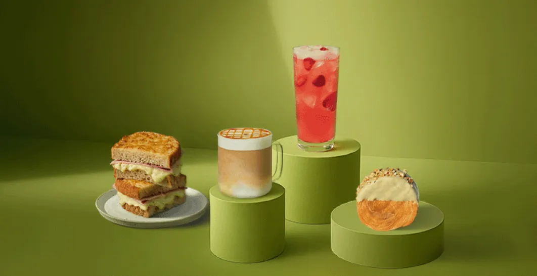 A photo of some of our favourite products - a vibrant red Strawberry Açai Refresha™, our classic hot caramel macchiato complete with cross-hatched drizzle on top, a signature grilled cheee toastie on a plate, and a delicious Pistachio scroll pastry, all on a green table.